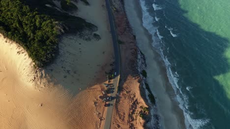 Tilt-up-aerial-drone-shot-revealing-the-stunning-Cliffs-of-Cacimbinhas-in-Pipa,-Brazil-Rio-Grande-do-Norte-with-tourists-sand-boarding,-a-paraglider-flying-around,-and-waves-crash-into-the-beach