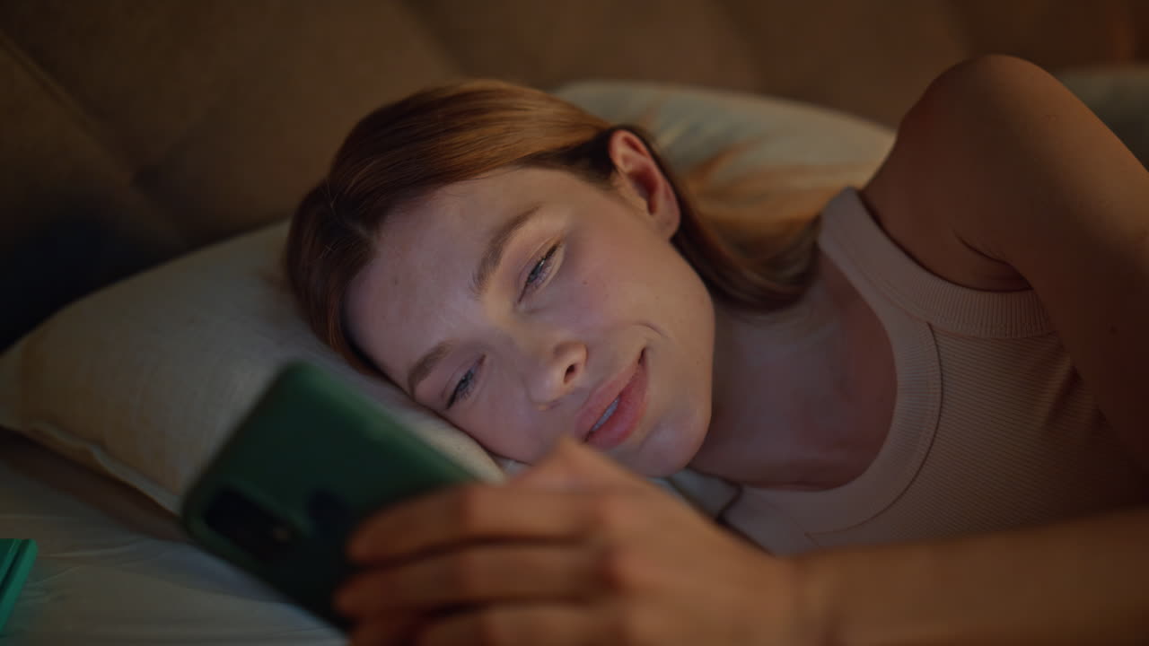 Evening woman looking smartphone on bed pillows close up. Smiling lady browsing