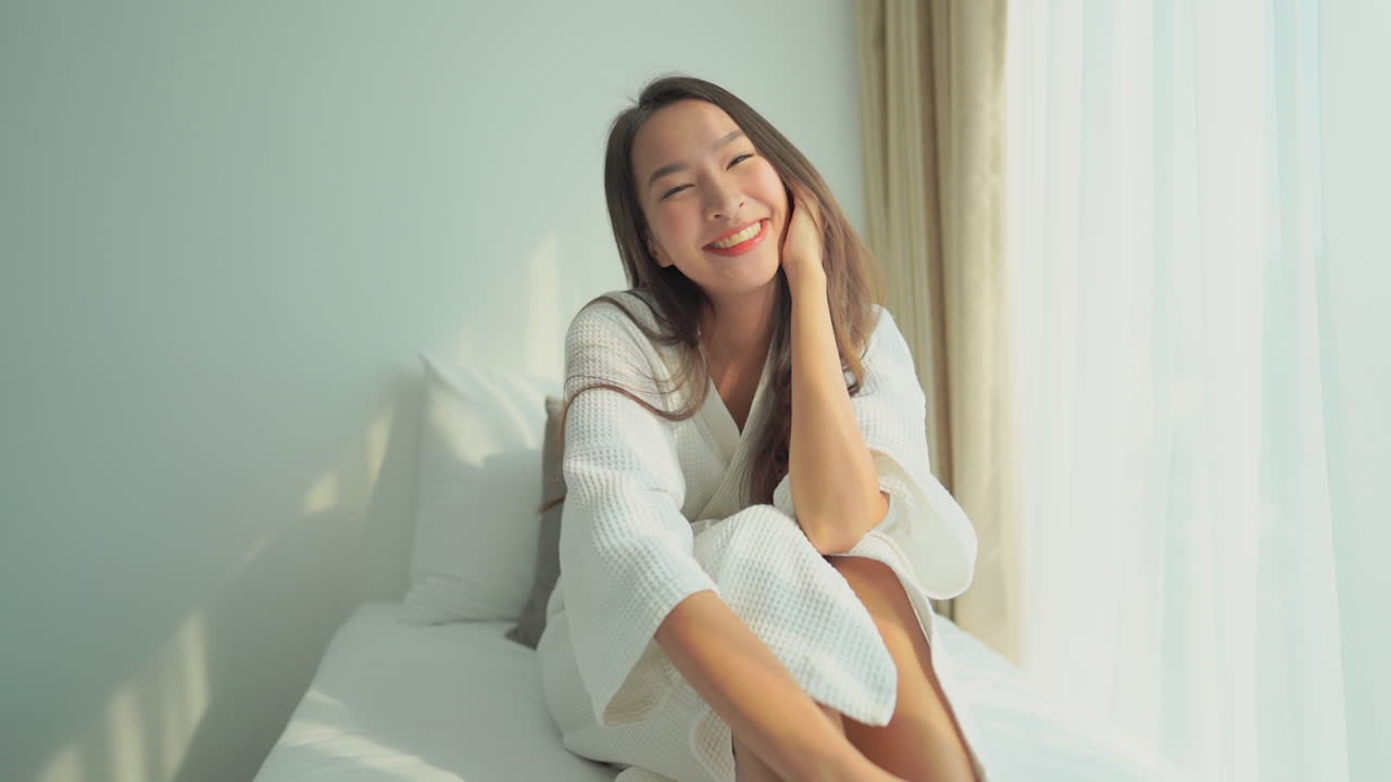 A young Asian woman dressed in a hotel robe, sitting in the window seat, enjoying the morning sun. Copy space