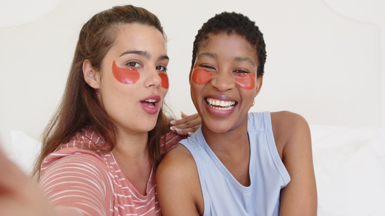 Two women friends sitting on bed left woman reaching phone demonstrating gel patches showing result