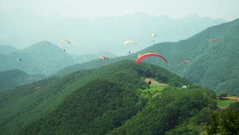 Gleitschirm,-Der-Von-Der-Spitze-Des-Hügels-Abhebt-Und-Auf-Viele-Andere-Menschen-Zufliegt,-Die-Um-Grüne-Berge-In-Der-Stadt-Danyang,-Südkorea,-Herumfliegen
