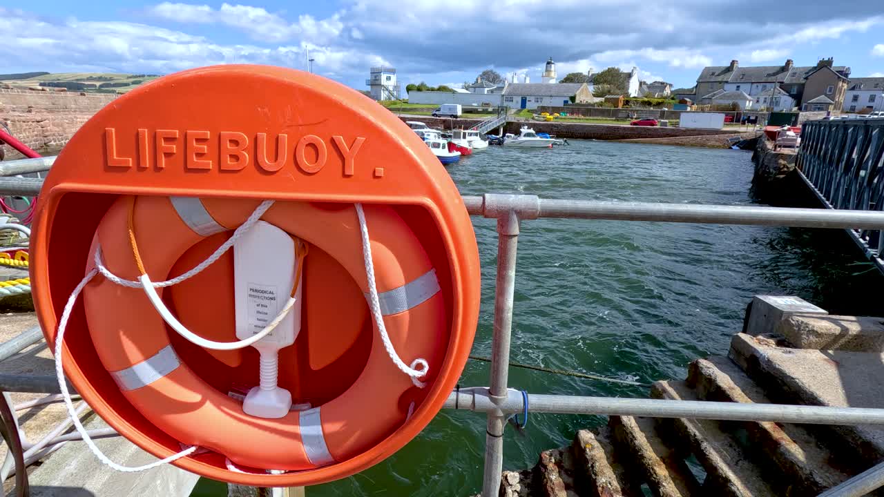 A bright orange lifebuoy ring mounted on a pier railing, with choppy green water and a coastal Scottish town in the background under daylight. Slight camera movement reveals more of the pier and water