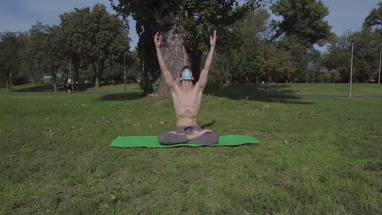 Joven deportista con mascarilla haciendo yoga en el parque sobre la esterilla verde, yogui realizando estiramientos de yoga en la naturaleza