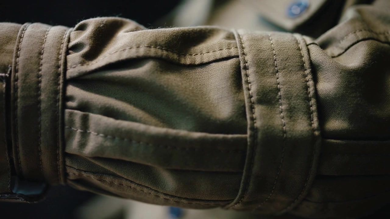 Frame opening showing forearm in olive jacket sleeve settling in dim space, with strap buckle