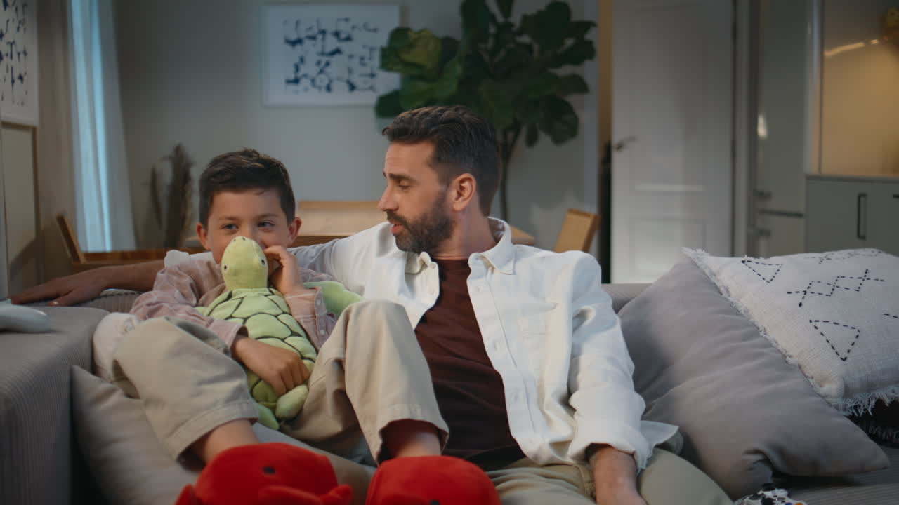 Father son talking evening sitting sofa at living room. Cute toddler holding toy