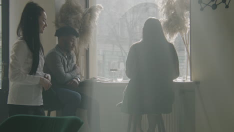 couple sit near window talk and drink wine african american man and asian woman sitting at the table near window talking and drinking wine female waiter brings delicious dishes to guests multi ethnic people in restaurant with modern design