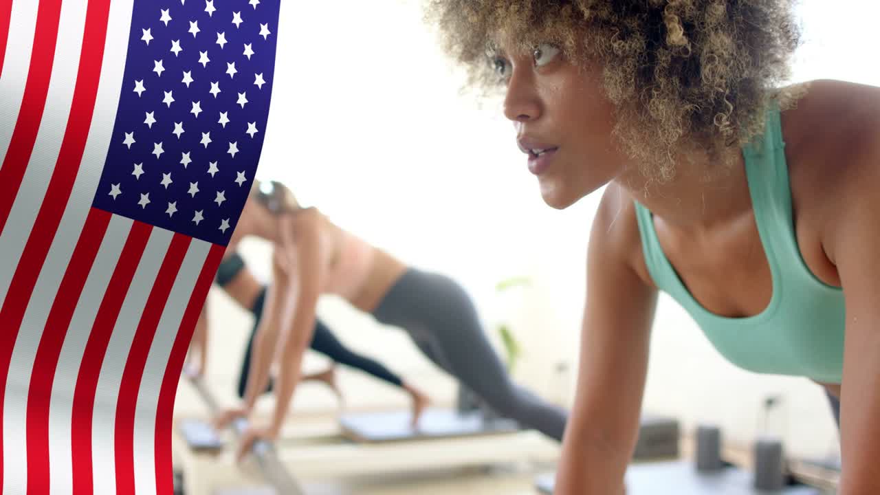 Frau vorne hält Plank auf Reformer, während Kamera verfolgt, Flaggen-Overlay verdeckt links, Fitness