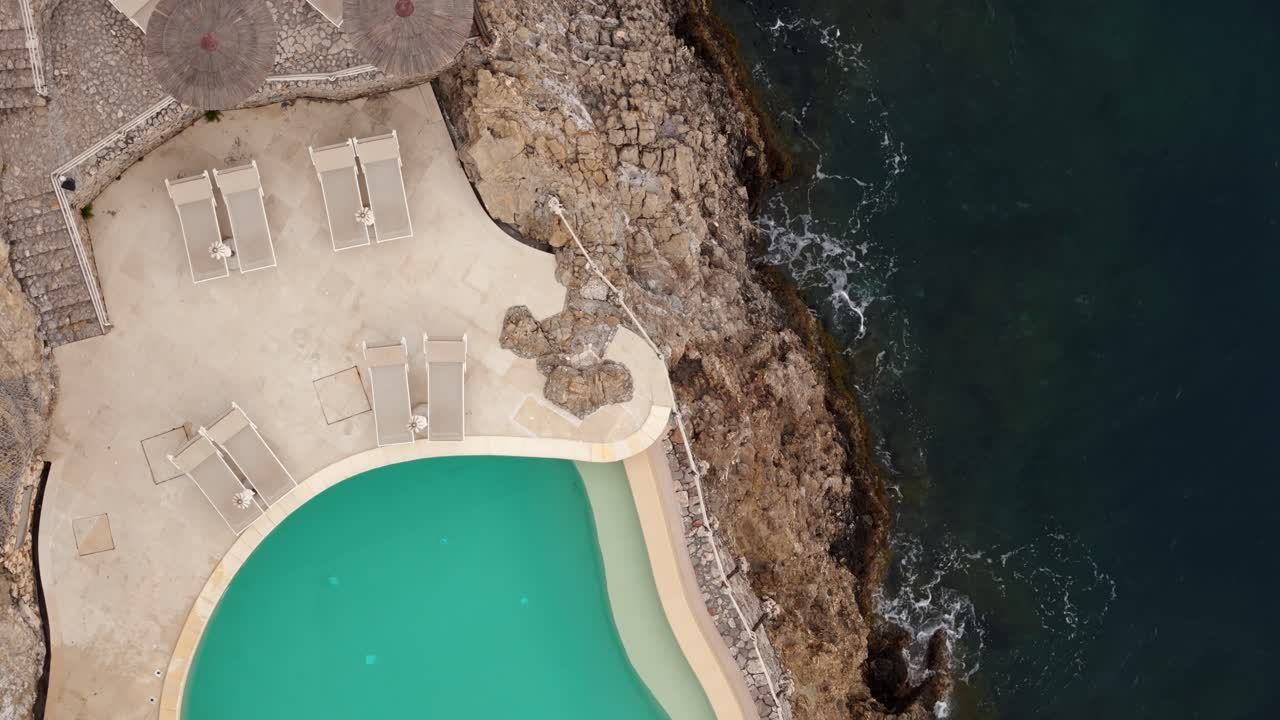 Aerial shot revealing the swimming pool at a fancy hotel, with waves crashing on the rocks, capturing a White Lotus vibe, in Taormina, Sicily, Italy (Sicilia, Italia)