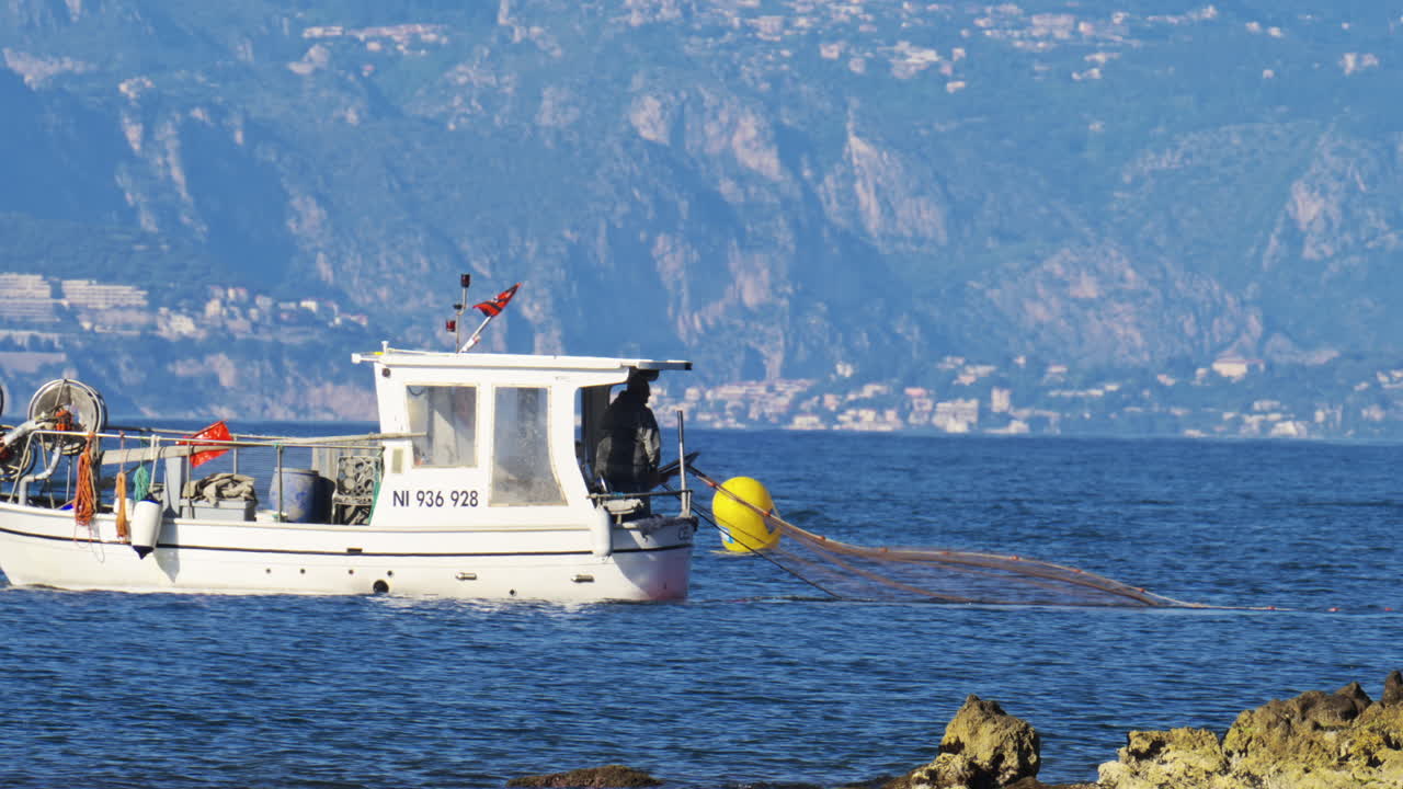 Antibes, France - May 2, 2025: View of a man in a white boat throwing a fishing net into the blue water in daylight