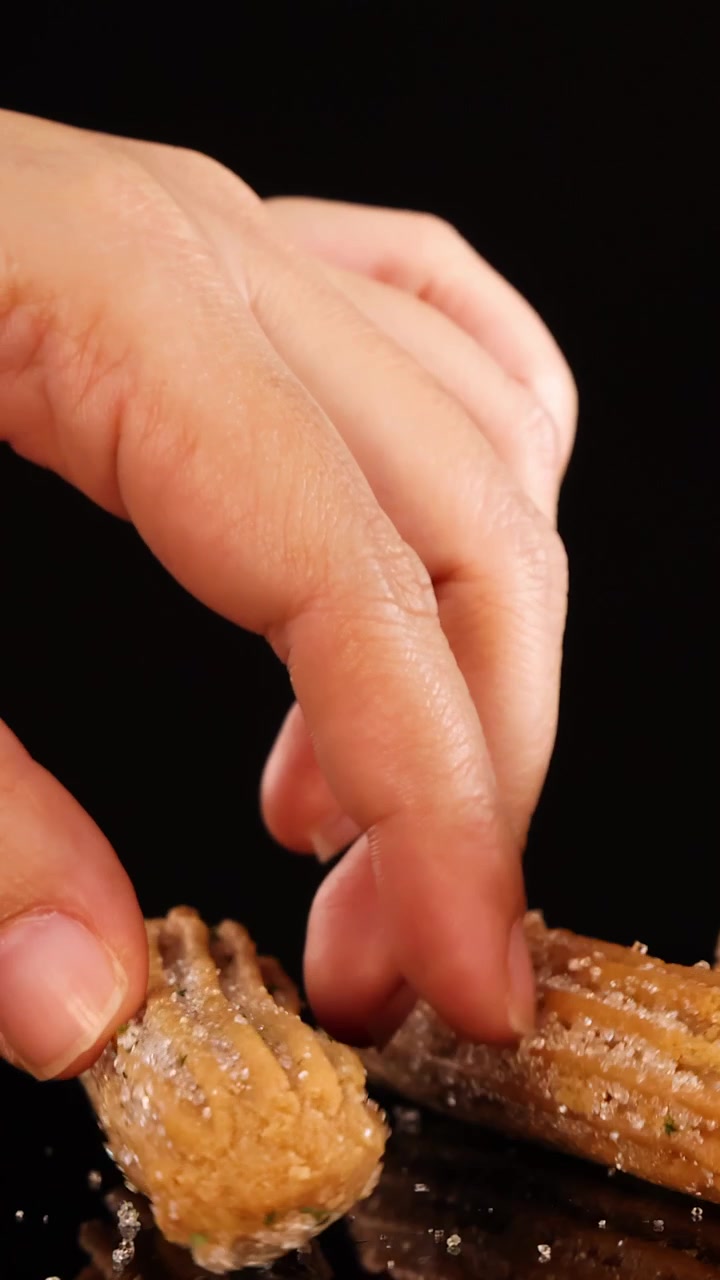Una mano solleva delicatamente un churro zuccherato da una pila ben ordinata, su uno sfondo nero con illuminazione da studio drammatica e inquadratura ravvicinata.
