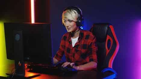 young woman with short blond hair in big headphones sitting in the gamer chair playing a game on the computer in a room with colorful neon lamps on the wall 1