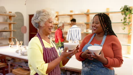 Happy-diverse-female-potters-holding-bowls-and-smiling-in-pottery-studio,-slow-motion