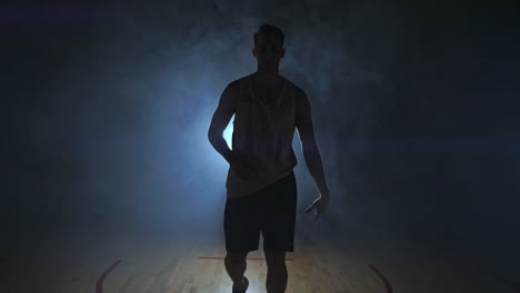 basketball player goes straight to the camera in a dark room with a backlit back in the smoke looking at the camera in slow motion. steadicam