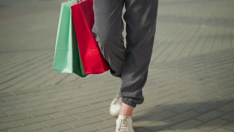 vista de la pierna de alguien con corredores grises y zapatos blancos de lona sosteniendo bolsas de compras verdes, menta y rojas mientras camina casualmente por un camino entrelazado, las bolsas se balancean con cada paso