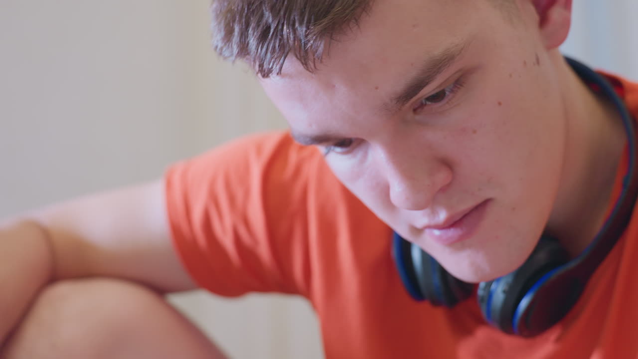 Close up of man wearing orange shirt and black headphones seated indoors holding screwdriver against chin while appearing thoughtful in moment of concentration reflection
