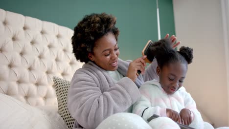 Happy-african-american-mother-combing-hair-of-daughter-using-tablet-in-bedroom,-slow-motion
