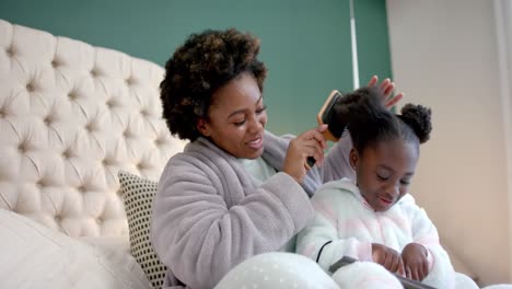 happy african american mother combing hair of daughter using tablet in bedroom, slow motion