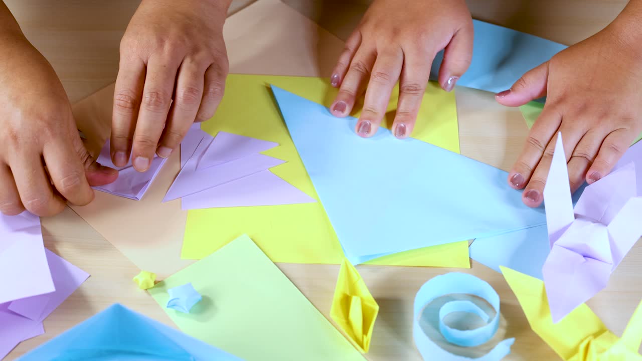 Multiple hands collaboratively folding pastel origami paper on a well-lit wooden table, overhead view