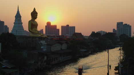 Longtail-Boat-on-a-river-in-Bangkok,-Thailand-with-the-sunset-and-big-golden-buddha