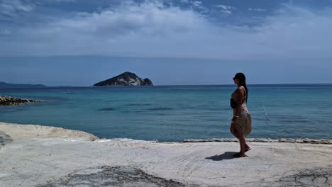 mujer caminando en una playa serena con una isla distante y un mar azul tranquilo bajo un cielo parcialmente nublado