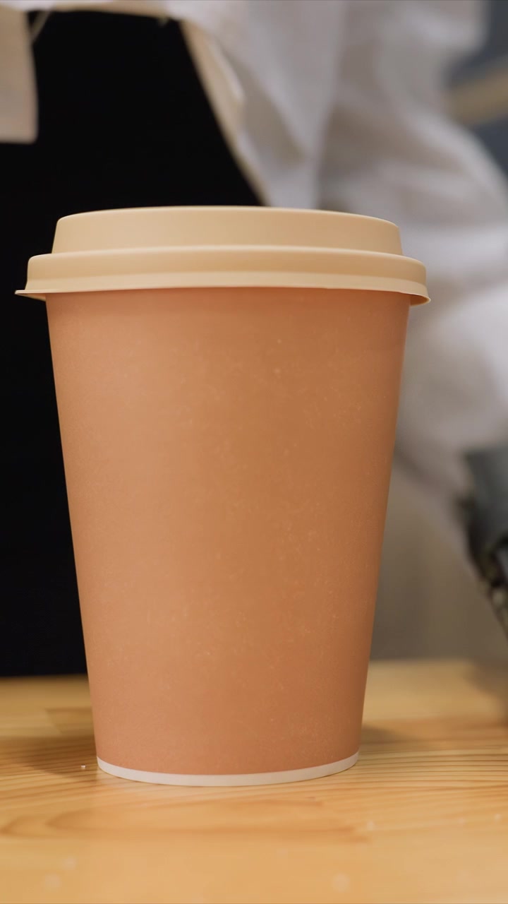 Primo piano di una tazza di caffè di carta marrone con un coperchio beige. La tazza rimane centrata mentre lo sfondo rimane leggermente sfocato. Un tavolo di legno presenta venature visibili sotto un'illuminazione costante