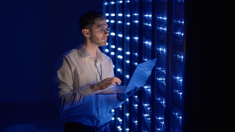 Male-network-engineer-doing-a-system-check-standing-in-the-server-room-with-his-laptop.-At-data-center-men-server-specialists-inspecting-working-system-and-hardware-of-rack-server-computer-cabinets