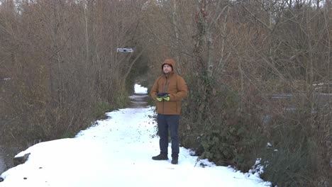 Warm-dressed-man-standing-on-snowy-pathway-controlling-drone-flying-over-Brooklands-lake,-Dartford
