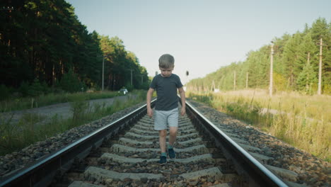boy in casual outfit walking slowly on rail track with green forest on both sides and blurred parked car visible in distance under clear sky on quiet peaceful day in rural setting