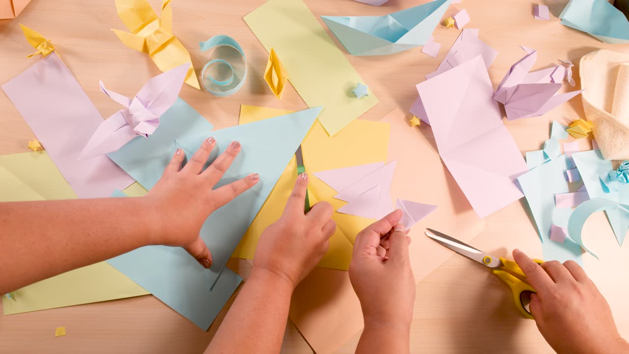 Two people cut and fold pastel paper for origami, overhead view, even soft lighting