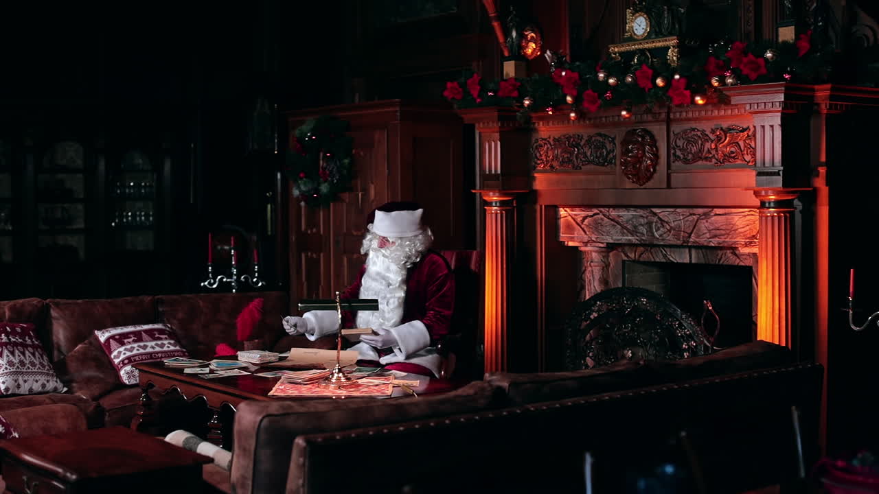 Beautiful cozy room with fireplace and old furniture. Santa Claus working sitting at the table looking through the cards.