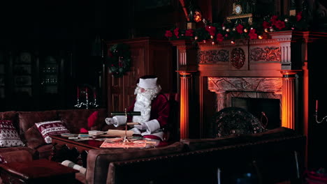 beautiful cozy room with fireplace and old furniture. santa claus working sitting at the table looking through the cards.