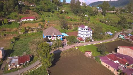 Aerial-view-of-a-village-in-Indonesia-surrounded-by-vegetable-farms,-trees,-fields,-volcanoes,-and-mountains-on-a-sunny-day