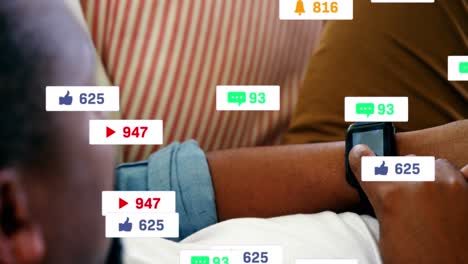 Animation-Von-Social-Media-Symbolen-über-Einem-Afroamerikanischen-Mann-Mit-Smartwatch,-Der-Zu-Hause-Auf-Der-Couch-Liegt