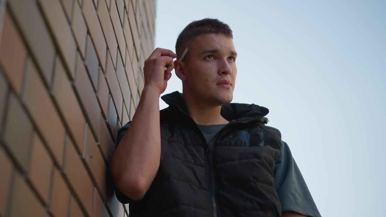upward view of young man resting against brick wall pulling cigarette from ear with focused expression as he prepares to light it in bright outdoor setting under clear sky