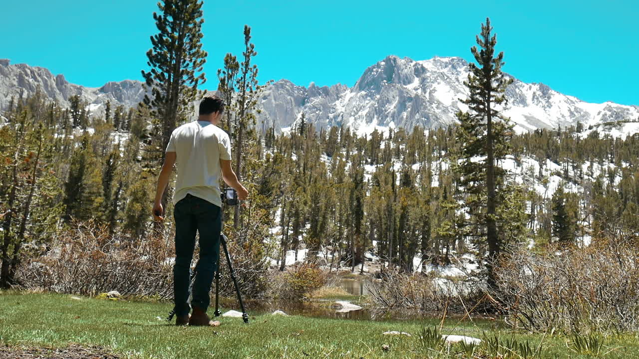 풍경 사진가가 눈 덮인 산봉우리를 배경으로 고산 호수 앞에 카메라를 설치합니다.