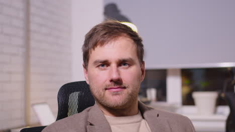 portrait of a smiling businessman sitting on office chair and looking at camera while two employees working behind him