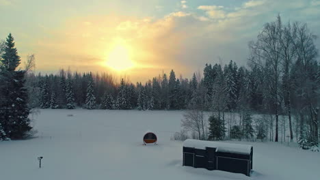 Drone-rising-backwards-over-a-distant-lakeside-Sauna-and-a-cottage,-wintet-sunset