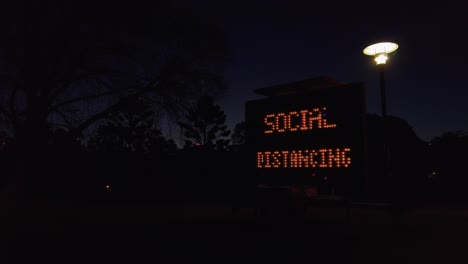 Social-distancing-electrical-sign-at-night-in-park-with-one-street-light,-slowmo