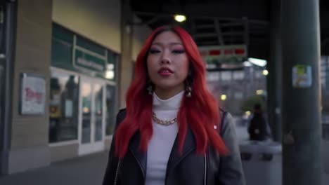 A young woman with striking red hair walks confidently through an urban area at twilight. Buildings surround her, and the atmosphere is lively with hints of nightlife.