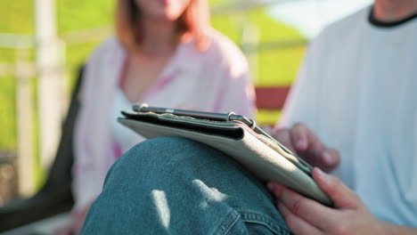 close-up of a man with leg crossed operating a tablet, a woman in pink slightly blurred in the background interacts and points at the screen, shifting focus back to her typing