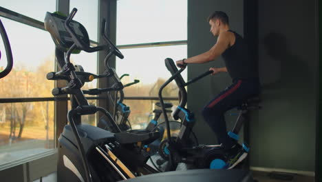 Fitte-Junge-Männer-Nutzen-Airbike-Im-Fitnessstudio.-Starker-Männlicher-Athlet-Beim-Cardio-Training-Auf-Dem-Fahrrad-Im-Fitnessstudio-An-Großen-Fenstern