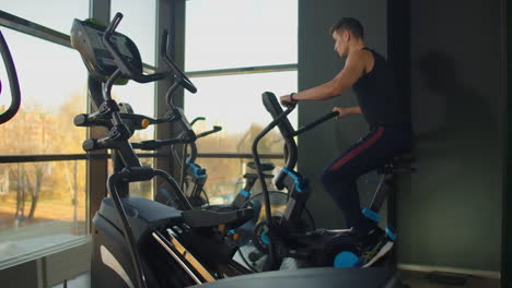 hombres jóvenes en forma usando bicicleta de aire en el gimnasio. atleta masculino fuerte haciendo entrenamiento cardio en bicicleta en el club de salud en grandes ventanas