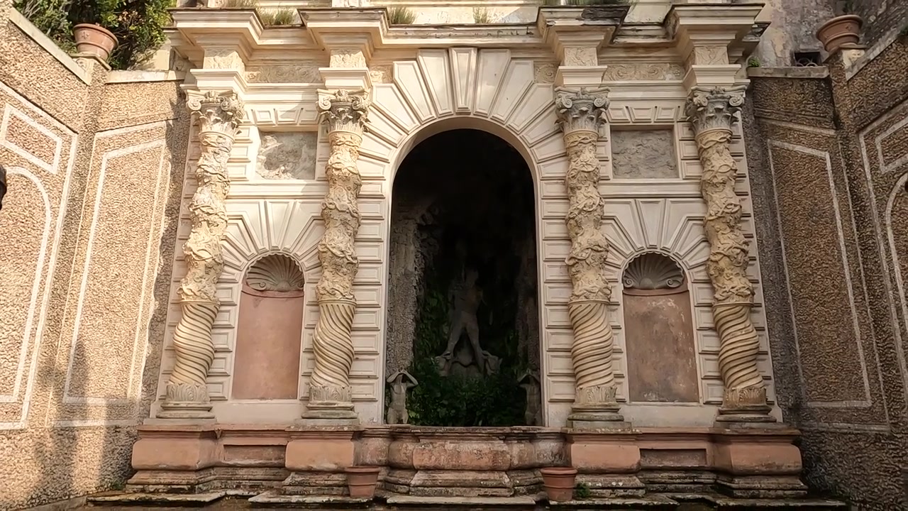 La telecamera si inclina all'indietro rivelando una nicchia di grotta barocca ornata con statua di figura classica, pareti ricoperte di edera e colonne di pietra intagliate