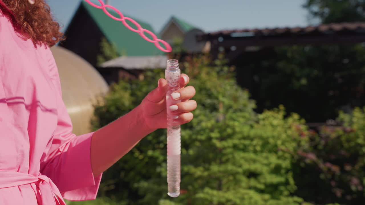 Caucasian Mother Holding Bubble Bottle Outdoors, Pink Dress Guiding Caucasian Daughter Along Garden Path, Closeup Of Wand And Solution, Warm Sunlight, Playful Mood, Candid Family Moment