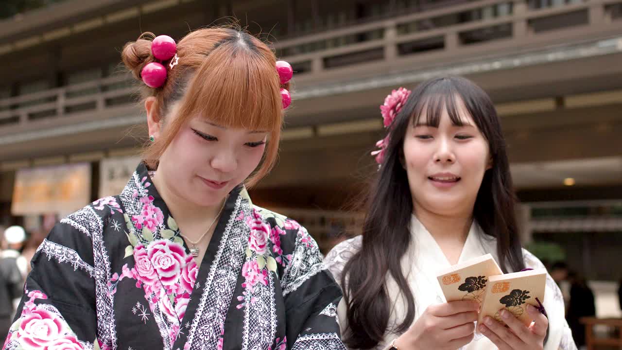 Çiçekli yukata giymiş iki gülen Japon kadın, bir Şinto tapınağında ahşap dua plaketleri tutuyor.