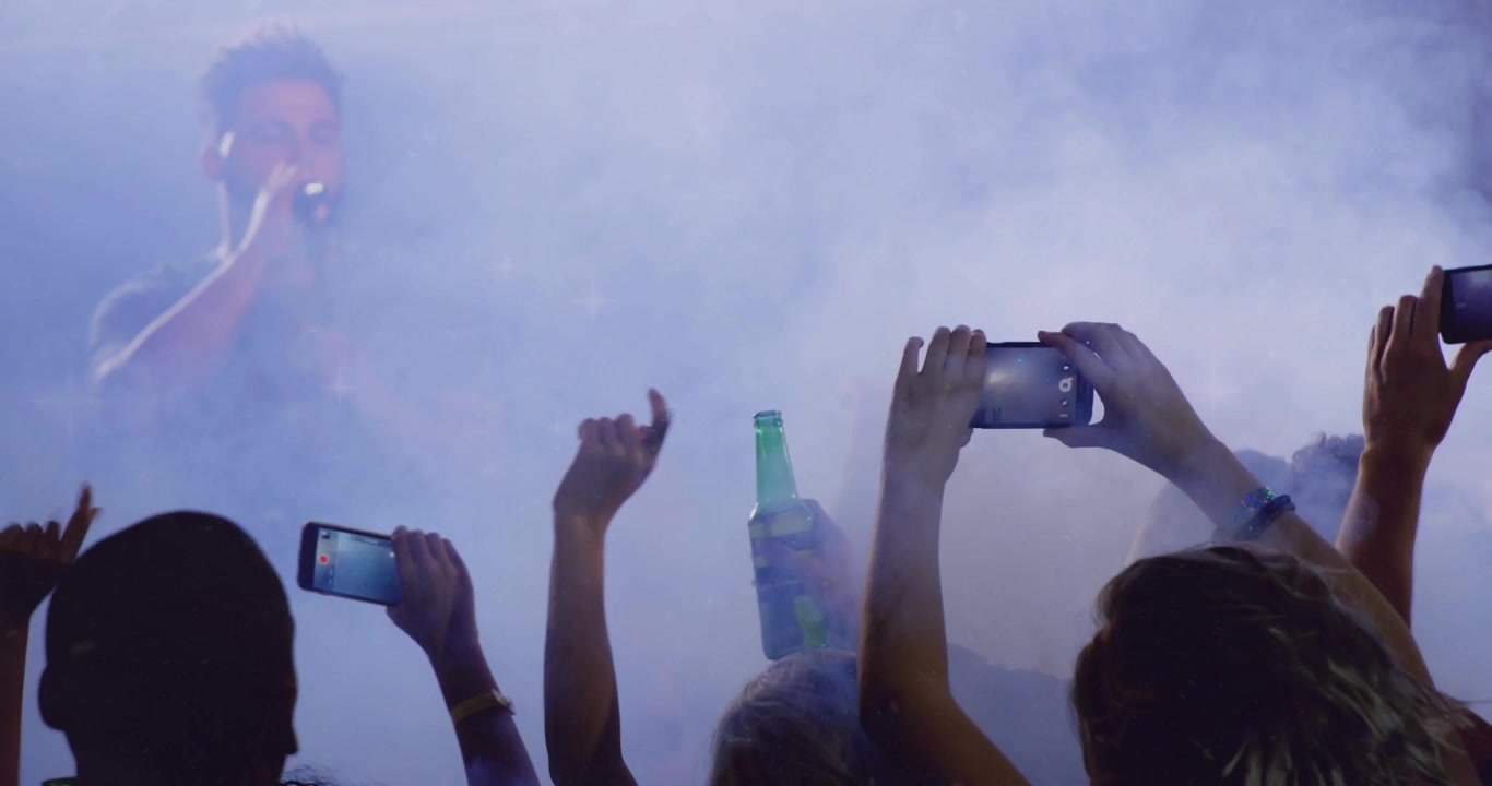Fans pressing stage raising phones and bottles recording music performance amid rainbow flare
