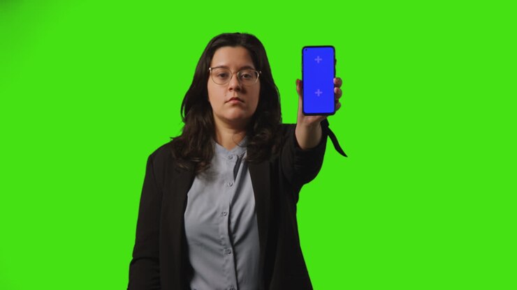 Giovane donna d'affari seria che tiene un telefono cellulare con schermo blu rivolto verso la telecamera, in piedi davanti a uno sfondo green screen