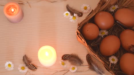 Spring-background-with-eggs-on-a-wicker-basket-with-some-hay,-feathers,-candles-and-daisies