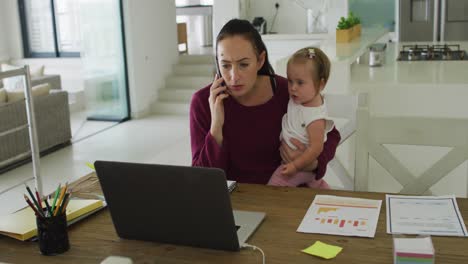 caucasian mother holding her baby and talking on smartphone while working from home