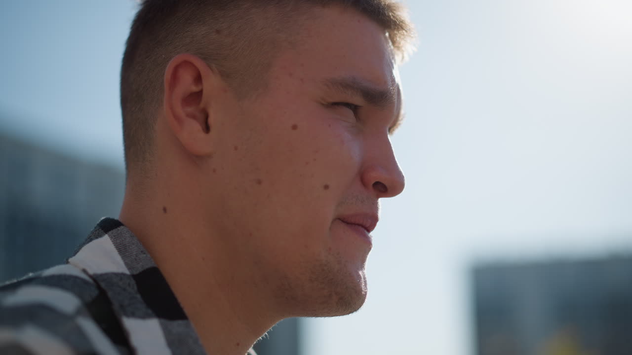Close up of man with visible facial spots taking white tablet while squinting in bright sunlight, with blurry background suggesting urban setting and blue sky during warm daytime